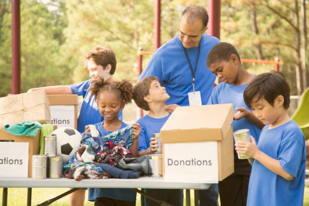 Multi-ethnic group of children on a soccer team work together to organize a charity drive for a local natural disaster event in their area.  The children set up a table at their local park to gather canned goods and clothing for the victims affected by the disaster.  Their team coach helps with the event.